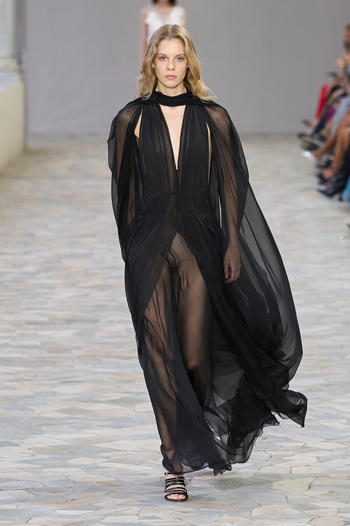 Runway model walking in a sheer black gown with cape-like sleeves and a plunging neckline at a fashion show.