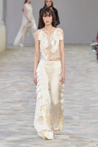 Model walks the runway in an ivory satin ensemble: sleeveless top with cascading ruffles and wide-leg pants with tiered frills.