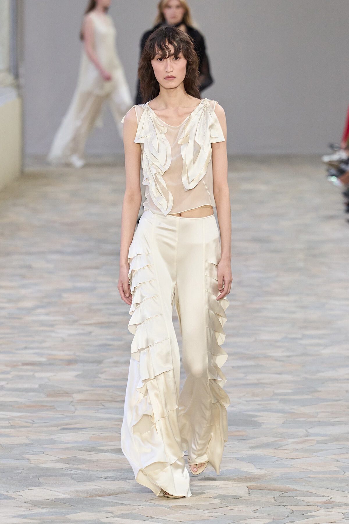 Model walks the runway in an ivory satin ensemble: sleeveless top with cascading ruffles and wide-leg pants with tiered frills.