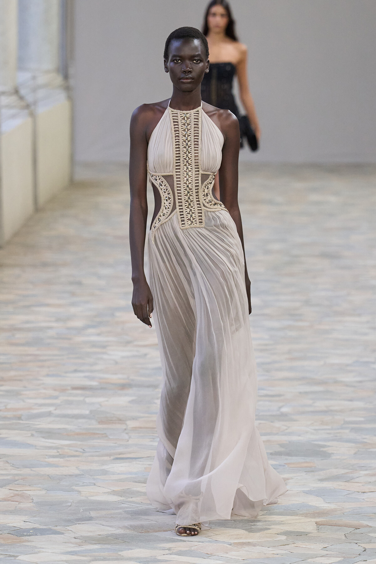 Model walks the runway in a pale beige halter gown with an ornate corset bodice and flowing pleated skirt.