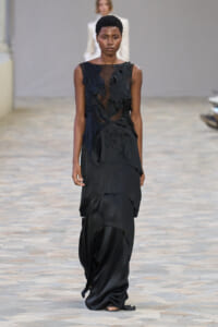 Black sleeveless gown with sheer bodice and layered, asymmetrical skirt on a runway.