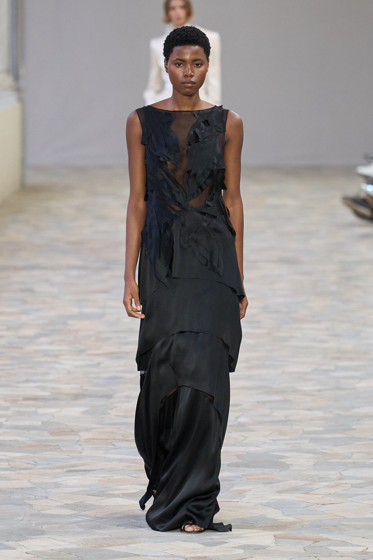 Black sleeveless gown with sheer bodice and layered, asymmetrical skirt on a runway.