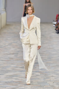 Model walks the runway in a cream satin tailored suit with a deep plunging neckline and a floral neck accessory.