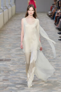 Model walks a runway in pale ivory satin ensemble: loose sleeveless tunic over wide-leg pants, with a sheer scarf streaming behind her as she moves.