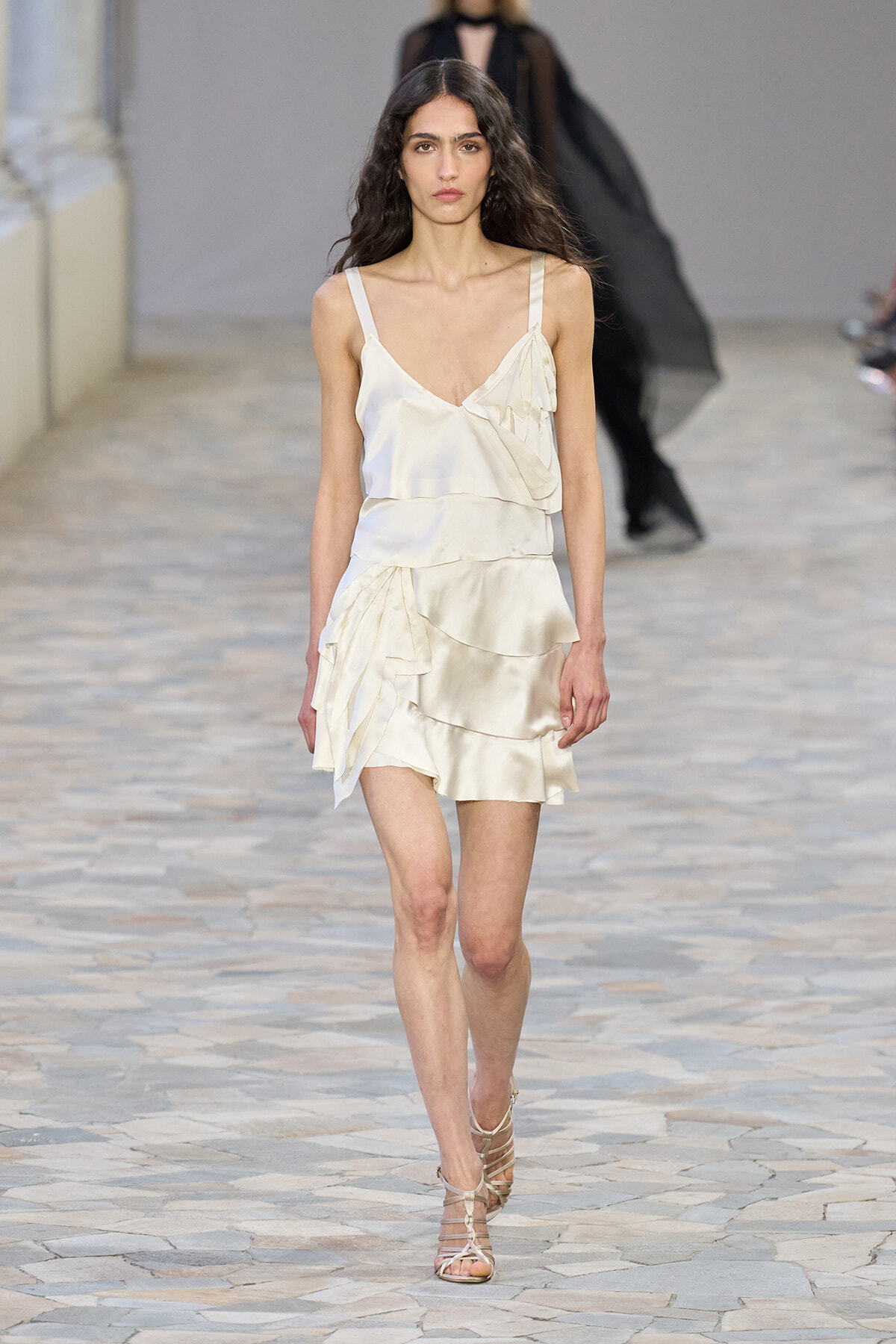 Female model wearing a cream satin, tiered dress with ruffled details and strappy sandals walking down a runway.