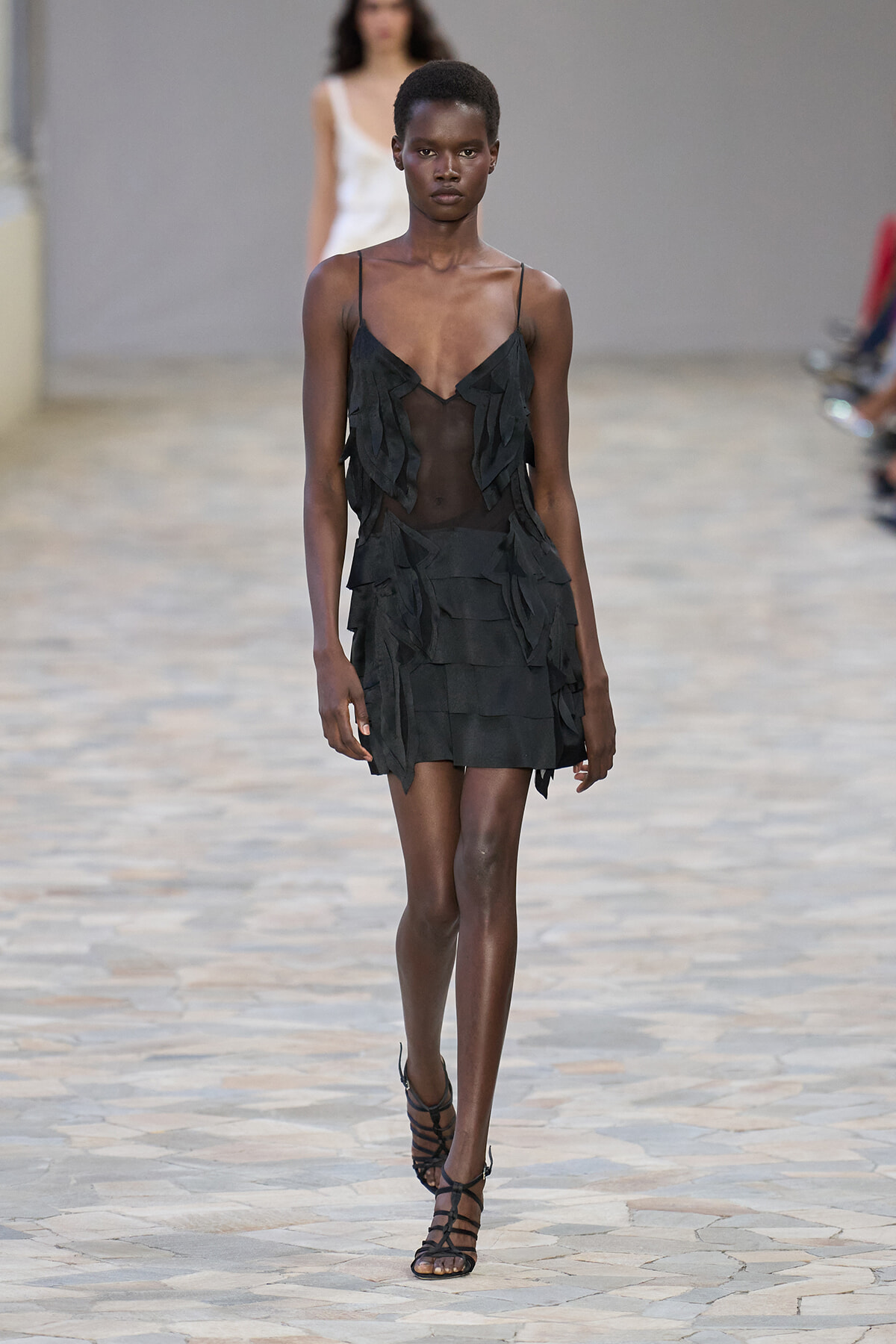 Model wearing a black fringed mini dress with sheer panels walking a runway.