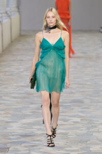 Model walking a runway in a teal sheer mini dress with layered ruffles and thin straps, carrying a dark green clutch.