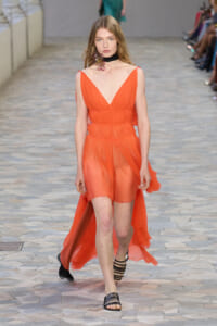 Model walking the runway in a bright orange, sheer chiffon dress with a deep V-neck and layered waist, paired with black strappy sandals and a black choker.