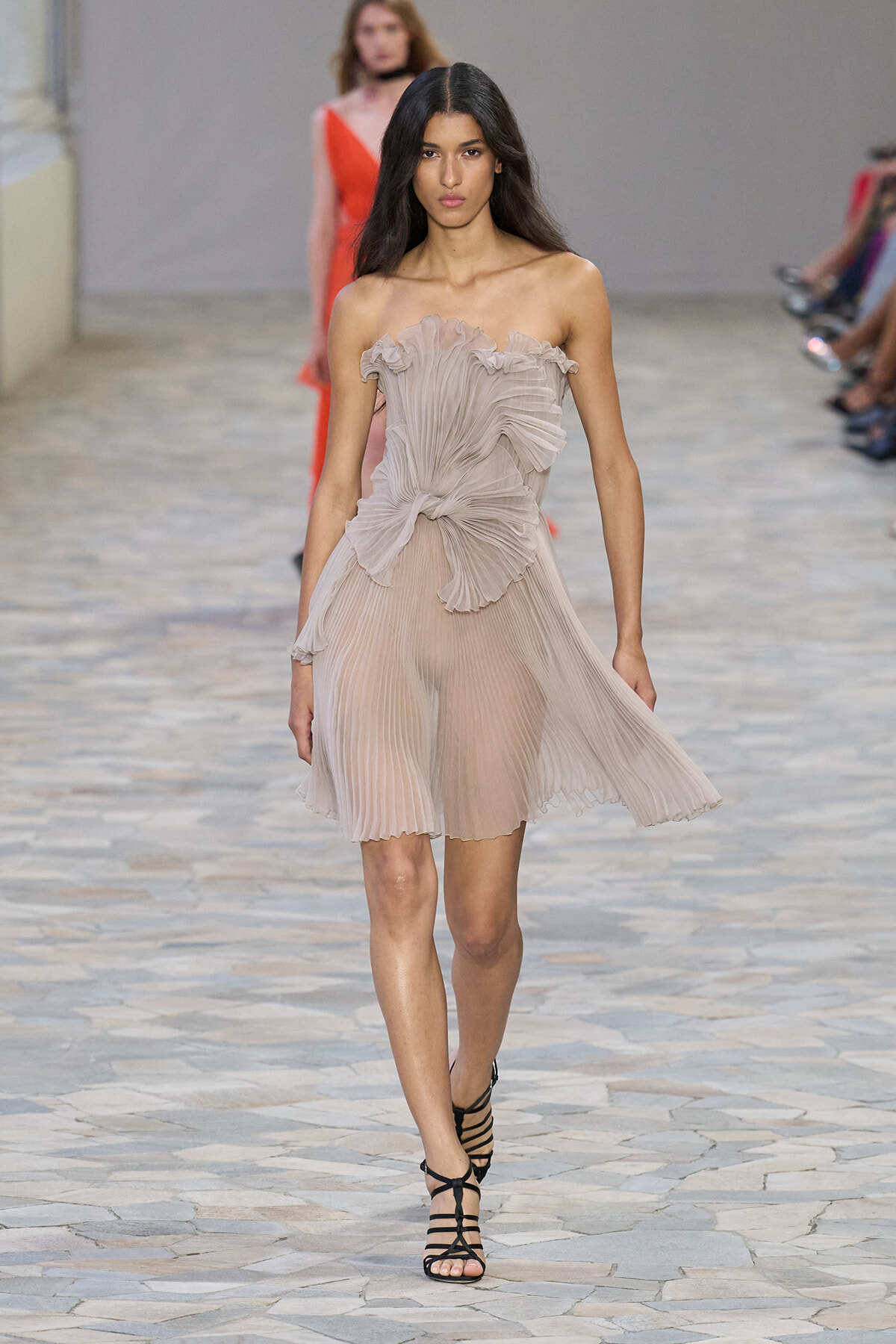 Model walking the runway in a beige pleated, off-shoulder dress with a knotted waist and black strappy sandals.