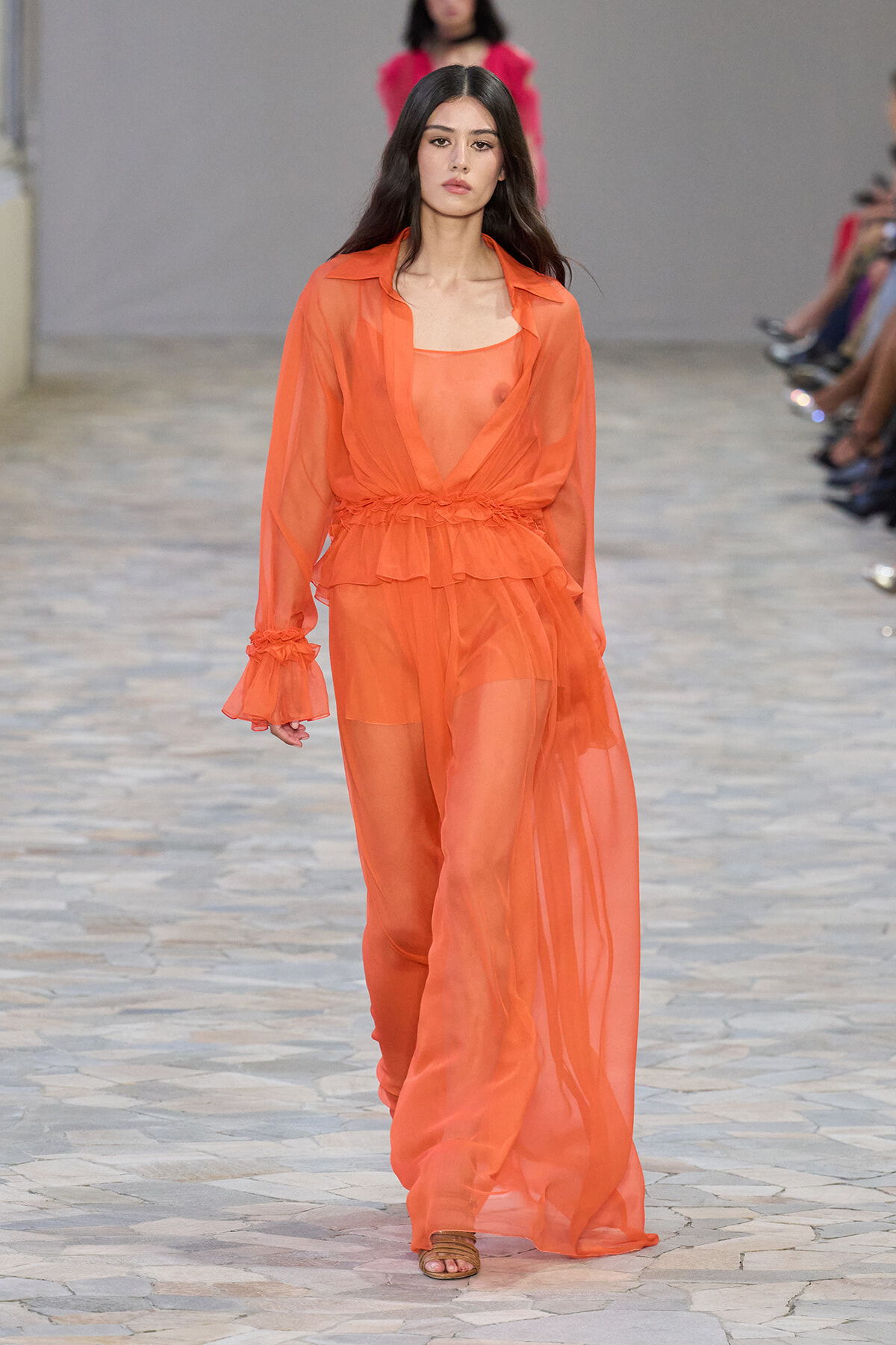 Model walks the runway wearing a sheer orange ensemble with a belted waist and flowing outer robe.