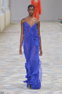 Female model walking a runway in a cobalt-blue, sheer, ruffled gown with thin straps and exposed shoulders.