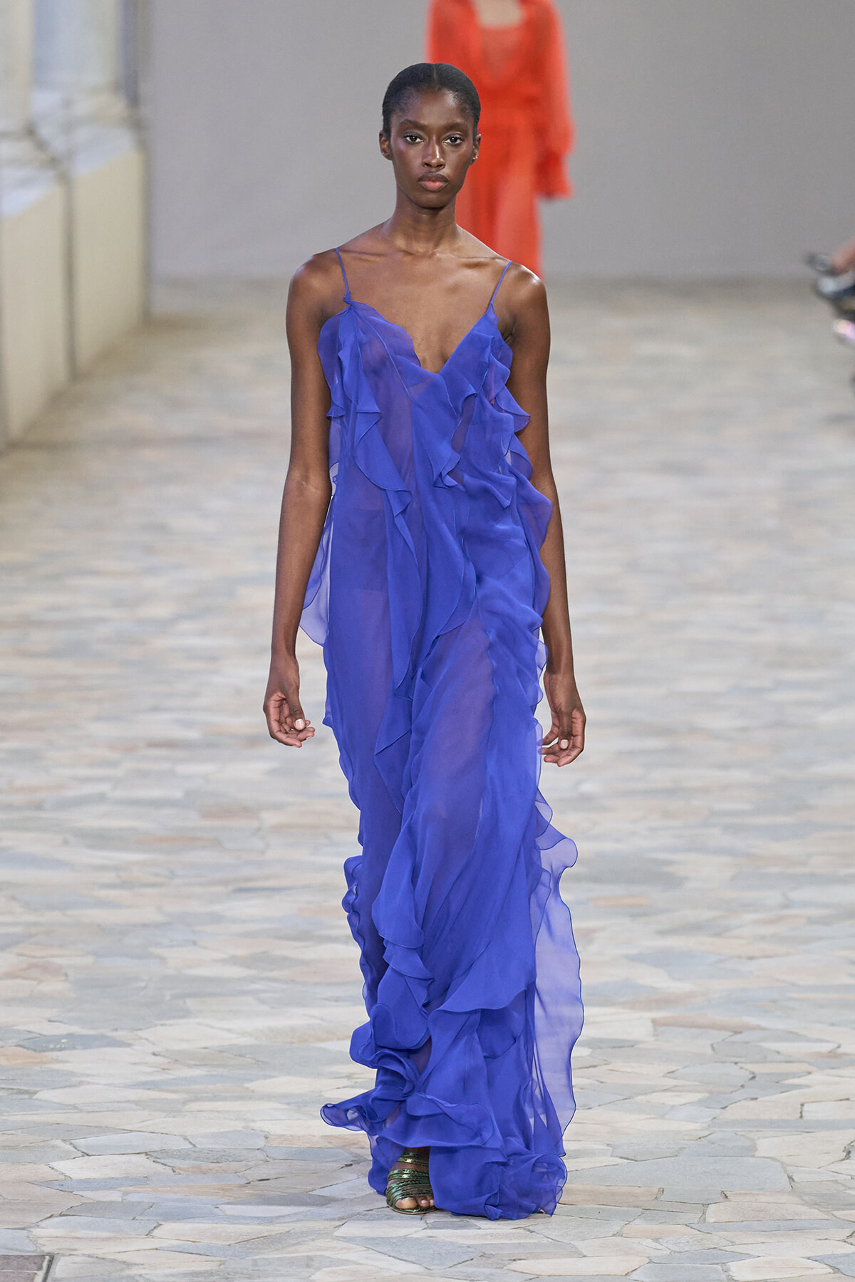 Female model walking a runway in a cobalt-blue, sheer, ruffled gown with thin straps and exposed shoulders.