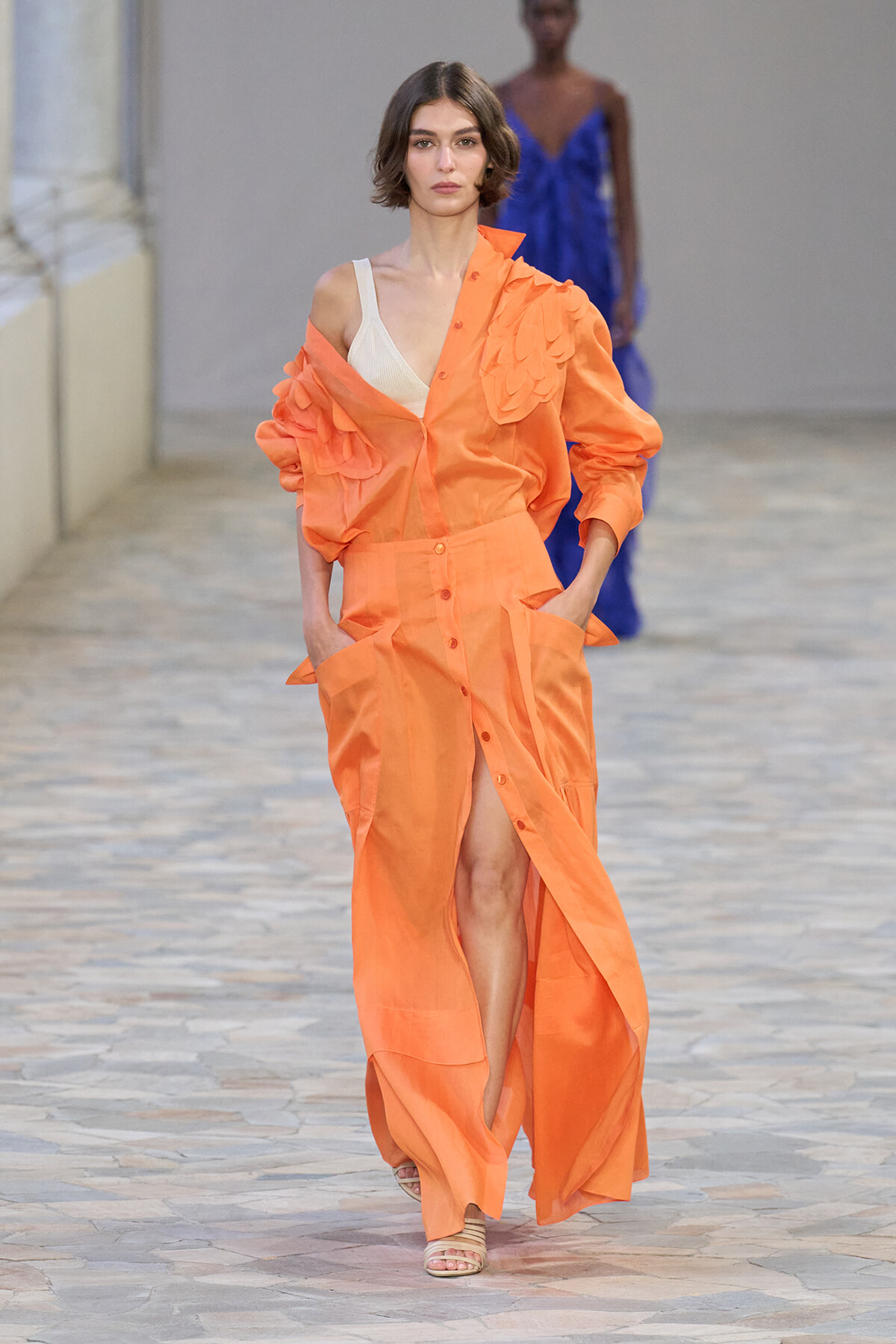 Model walks the runway in a bright orange coordinated outfit with voluminous ruffled sleeve and long buttoned skirt, beige top visible underneath.