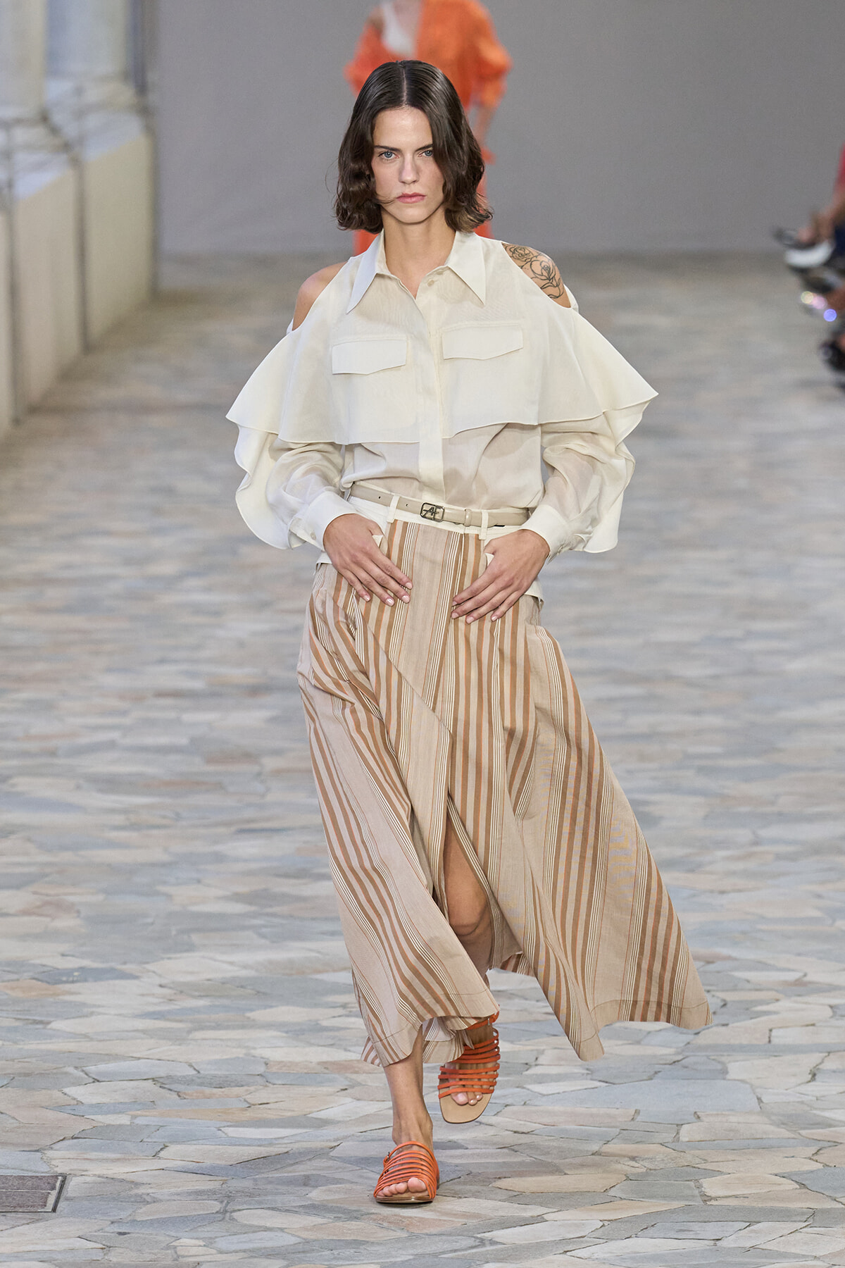 Runway model wearing a white cold-shoulder blouse with layered sleeves and a beige striped wide-leg skirt, plus orange sandals.