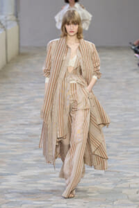 Model walks a beige striped layered suit and coat ensemble down a runway.