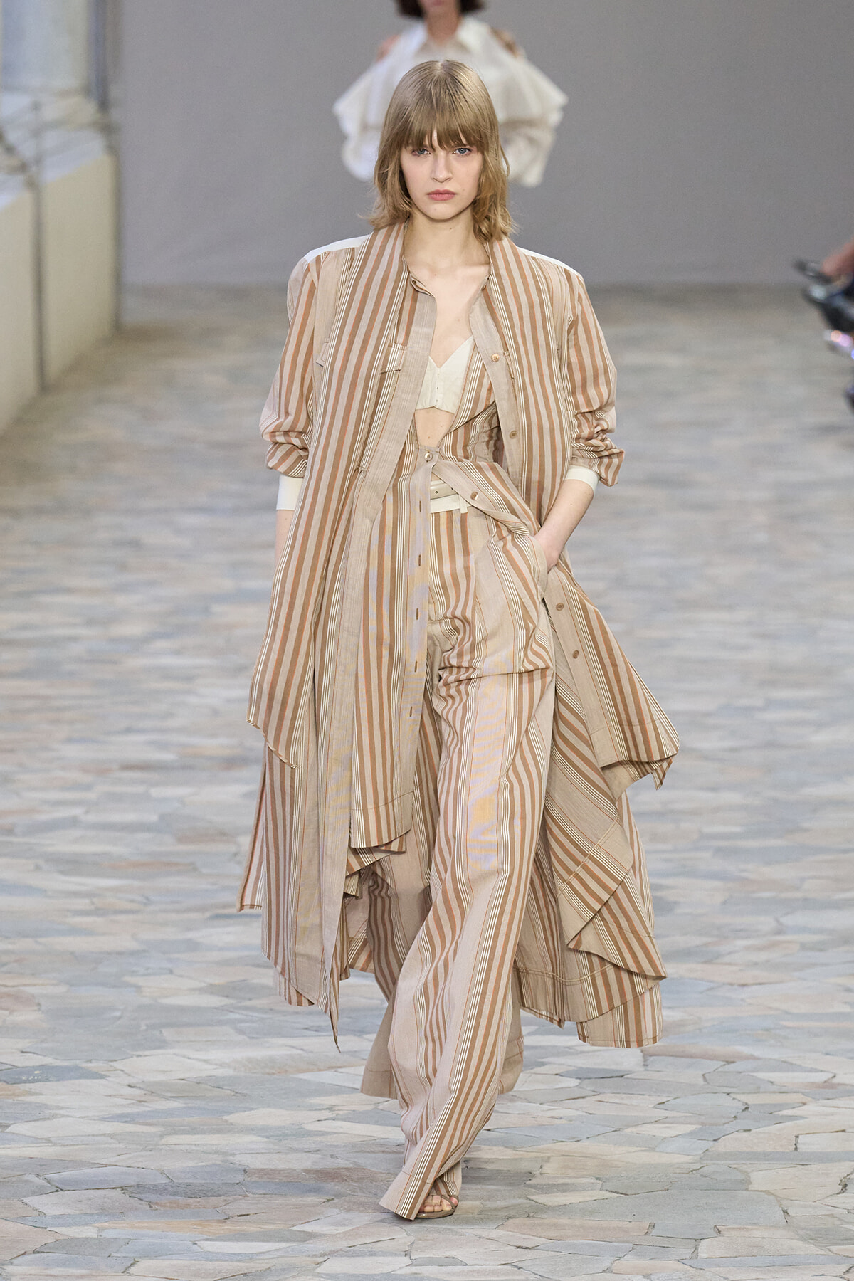 Model walks a beige striped layered suit and coat ensemble down a runway.