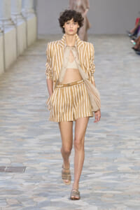 Model walking the runway in a beige striped blazer and matching shorts, with a light beige bralette and sandals.