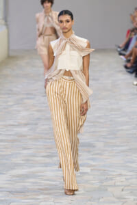 Model on a runway wearing a cream sleeveless top, striped scarf at the neck, and beige striped wide-leg trousers.