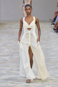 Model on runway in a white sleeveless cutout maxi dress with a plunging bodice and front buttons, walking forward with hands in pockets.