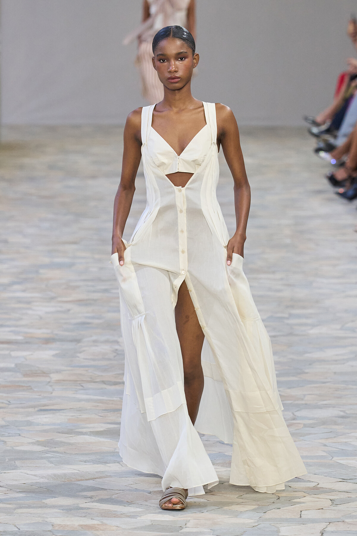 Model on runway in a white sleeveless cutout maxi dress with a plunging bodice and front buttons, walking forward with hands in pockets.