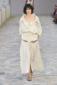 Model walks the runway in a cream, oversized hooded jacket over a matching bandeau top and wide-leg skirt; neutral sandals complete the look.