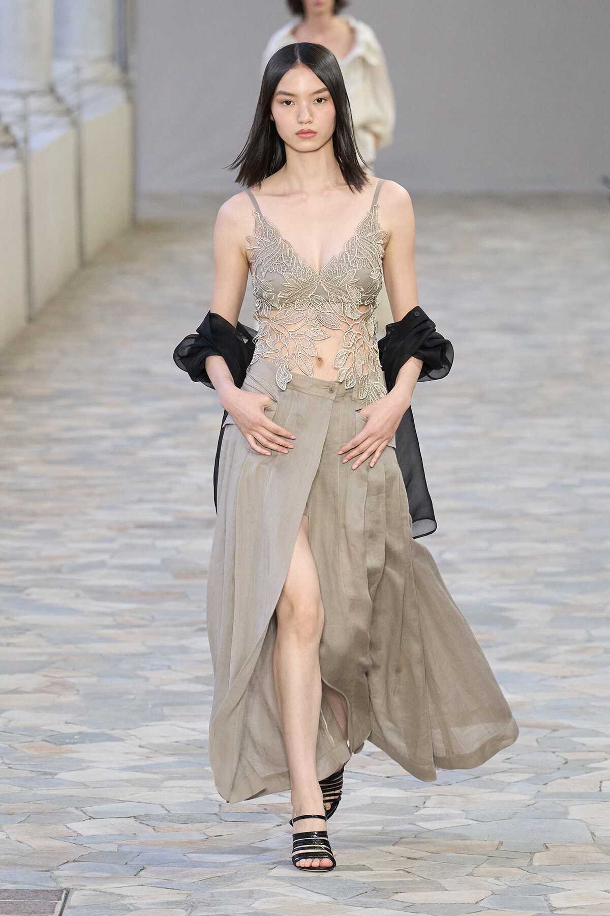 Model walking runway in taupe lace bodice top and high-slit beige skirt with a black sheer jacket over the shoulders.