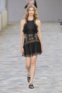 Model walking a runway in a black lace mini dress with sheer panels and scalloped hem, paired with strappy black sandals.
