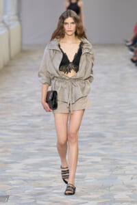 Model walks a fashion runway in a beige belted jacket with matching shorts, black lace bralette visible, carrying a black clutch.