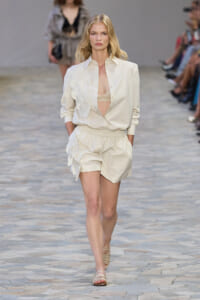 Model walking a runway in an ivory shirt with layered leaf appliqués and matching elastic-waist shorts; beige undergarment shows at the chest.
