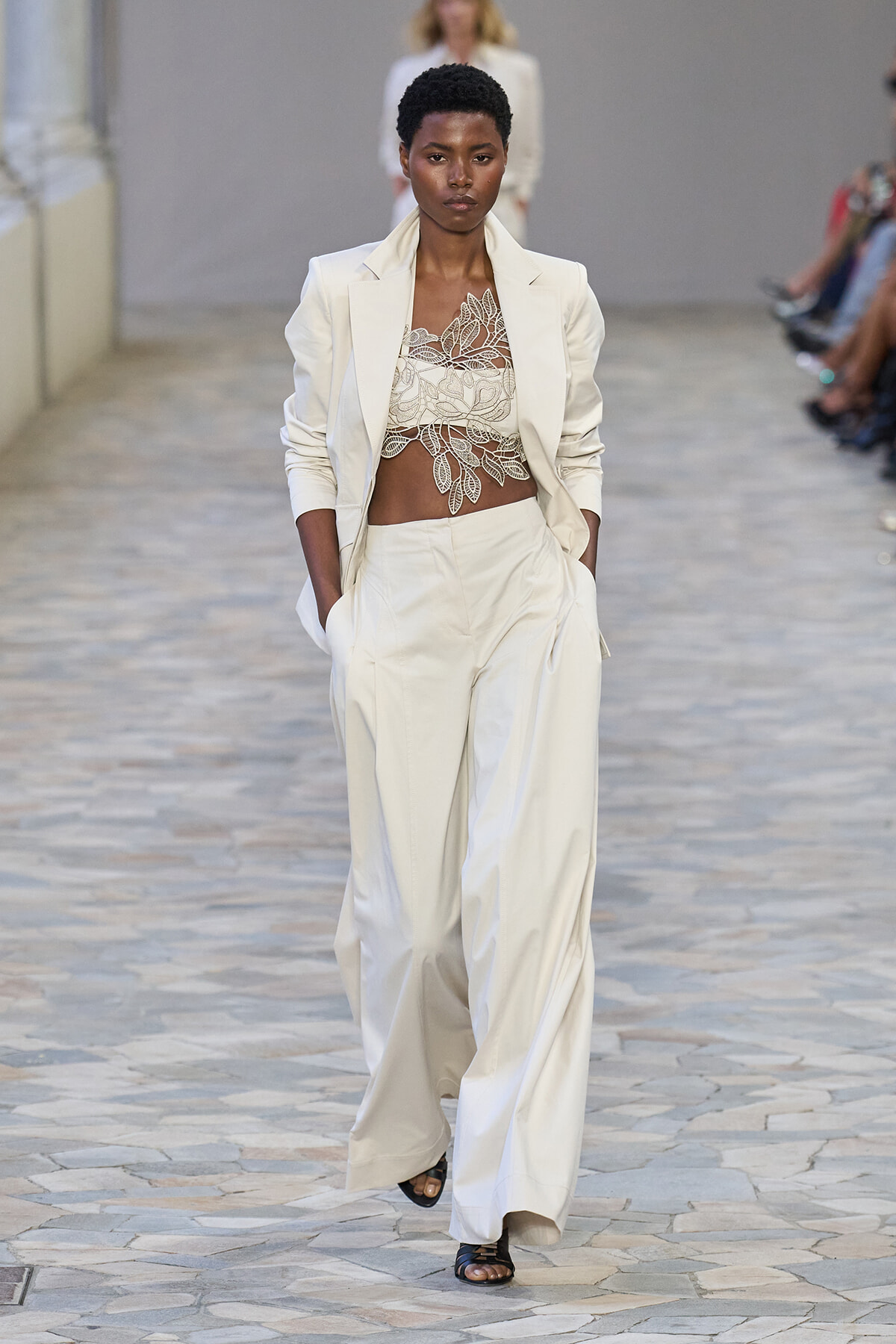 Model walking a fashion runway in a white suit with a lace crop top, hands in pockets.