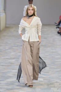 Model walking a runway in an off-shoulder ivory blouse with a structured peplum, paired with wide-leg beige trousers and a flowing black sheer cape; black floral choker accessory.