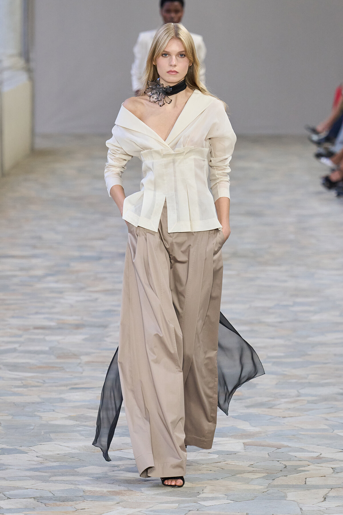 Model walking a runway in an off-shoulder ivory blouse with a structured peplum, paired with wide-leg beige trousers and a flowing black sheer cape; black floral choker accessory.