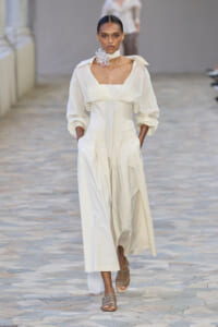 Model walks a runway in a cream, long-sleeved, wide-leg dress with structured bodice and a white floral neck accessory.