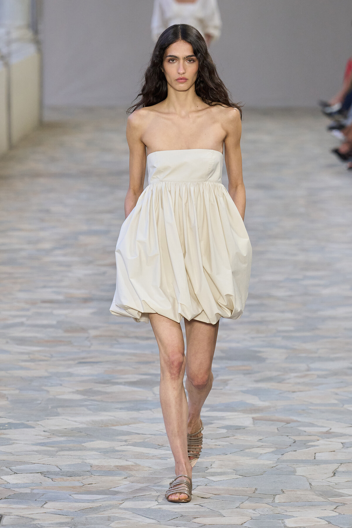 Female model walks a runway in a cream strapless bubble dress and flat sandals on a stone-tiled floor.