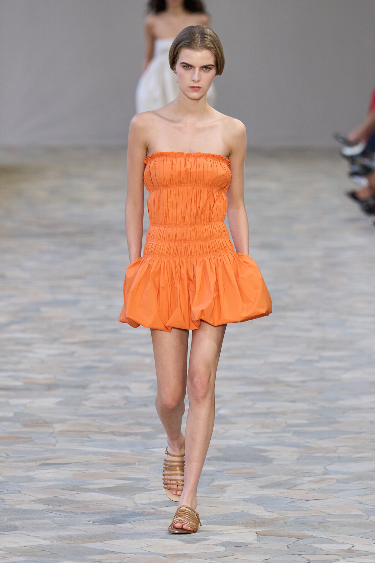 Model walking the runway in an orange strapless ruched mini dress with a voluminous skirt and brown strappy sandals.