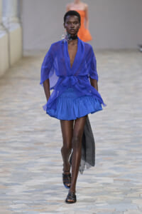 Model walking a fashion runway in a cobalt blue sheer wrap blouse tied at the waist and a matching ruffled mini skirt with black sandals and a floral choker.