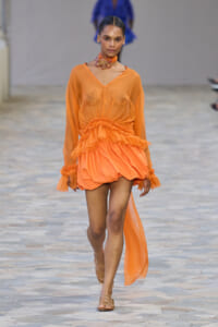 Runway model in a sheer orange dress with ruffled waist and long sleeves, wearing a floral neckpiece, walking the catwalk.