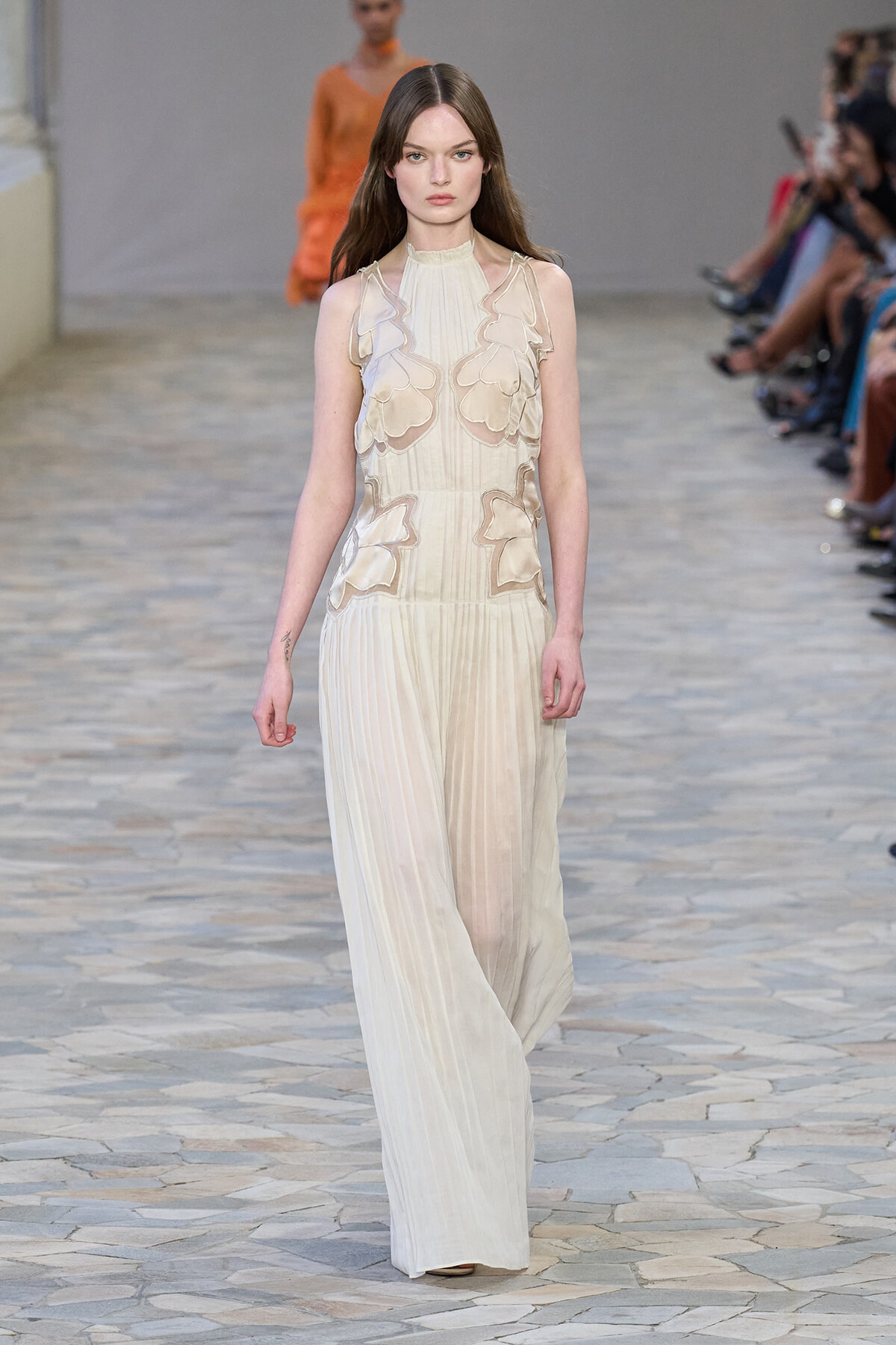 Model walks a cream pleated jumpsuit with sheer floral appliqués on the bodice, on a fashion runway with spectators