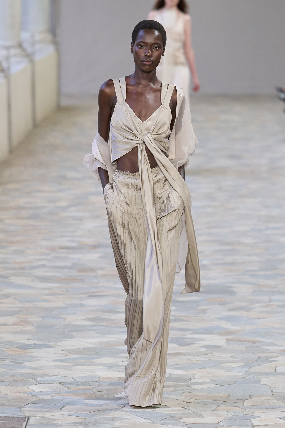 Model walking the runway in a beige satin twist-front blouse paired with wide-leg pleated pants.
