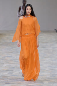 Model walking the runway in an orange sheer off-shoulder top with matching flowing pants and a jeweled neckpiece on the catwalk.