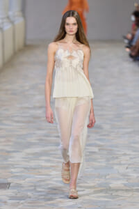 Model walks the runway in a sheer ivory cream top with floral appliqué and a matching pleated skirt with strappy sandals.