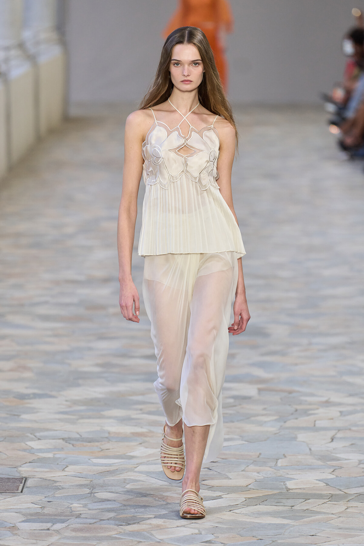 Model walks the runway in a sheer ivory cream top with floral appliqué and a matching pleated skirt with strappy sandals.