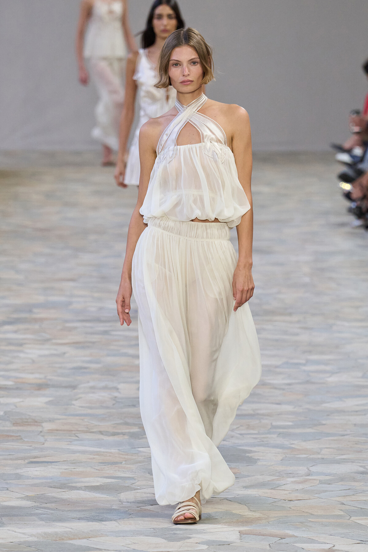 Model walking a runway in a white, sheer halter-neck dress with crisscross straps at the chest and a flowing skirt. sandals visible.