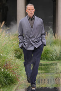 Man in a gray double-breasted blazer and dark trousers walking along a reflective water feature with tall grasses on either side.