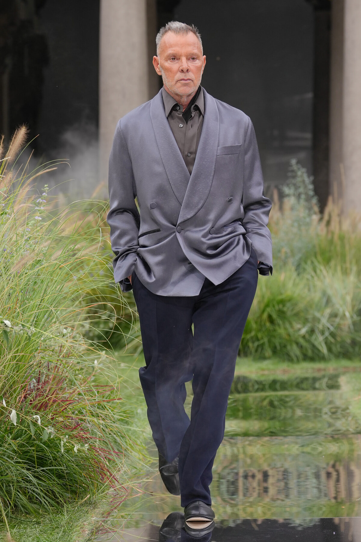 Man in a gray double-breasted blazer and dark trousers walking along a reflective water feature with tall grasses on either side.