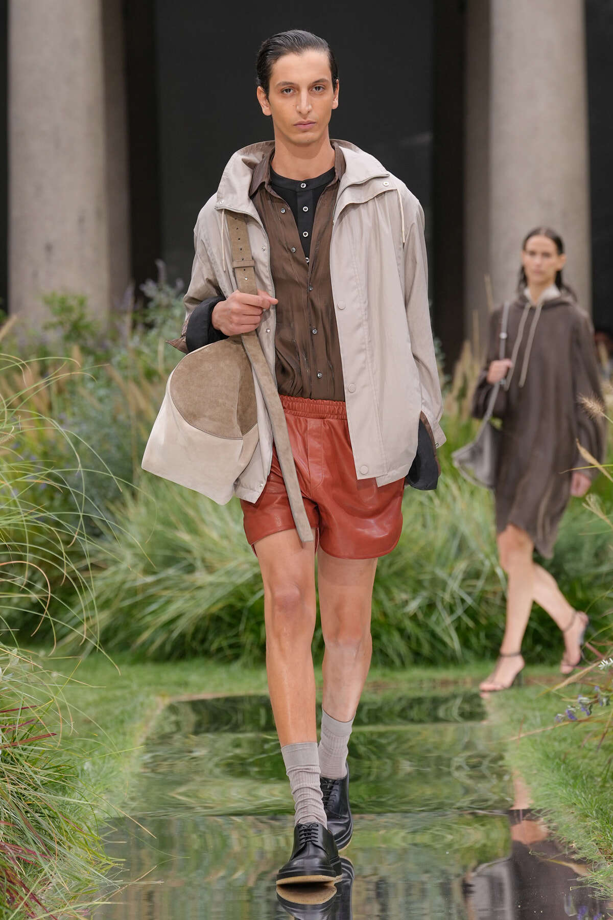 Male model walking a runway outdoors wearing a beige windbreaker, brown shirt, red leather shorts, gray socks, black shoes with a tan sole, and carrying a large beige bag over the shoulder.
