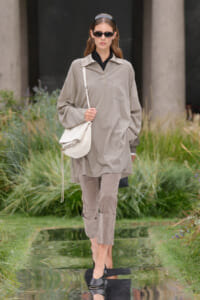 Model walking in a beige oversized shirt and matching pants, carrying a white shoulder bag outdoors, wearing sunglasses and headband.