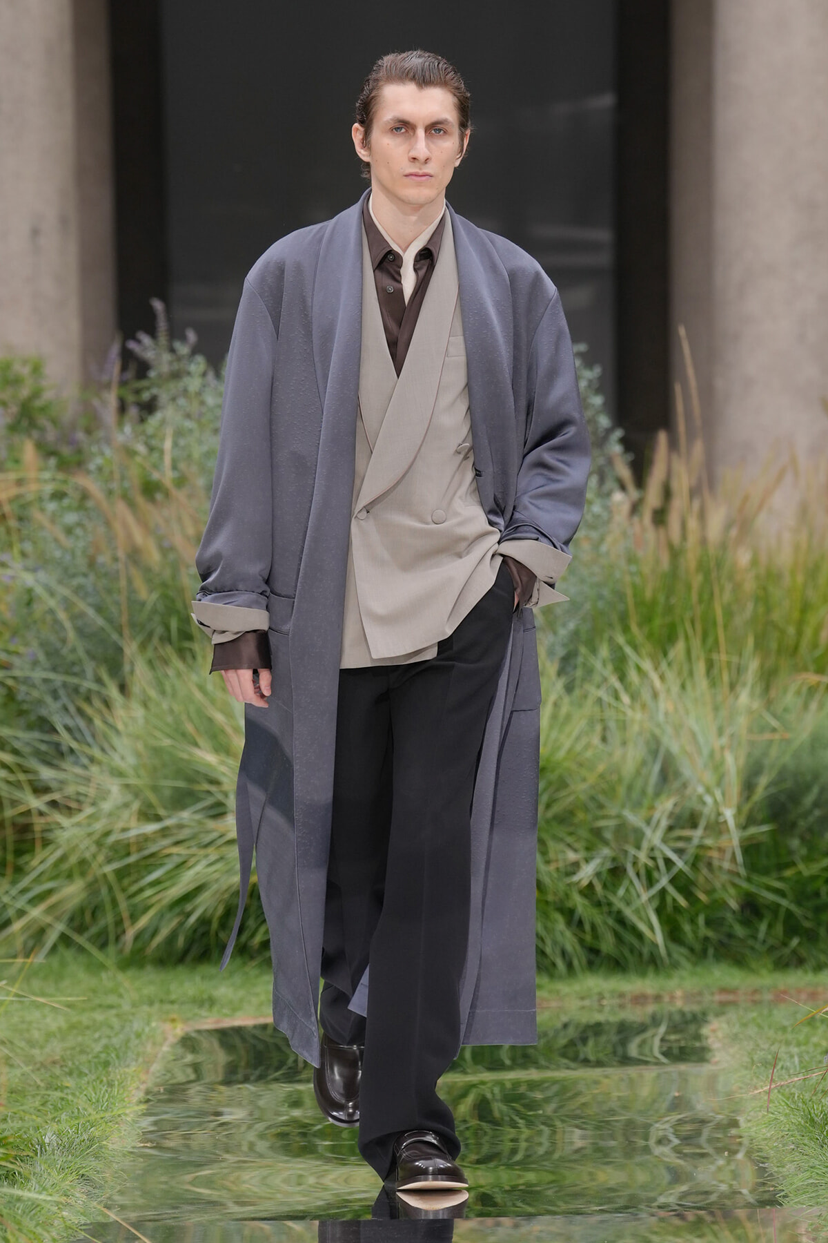 Male model wearing a long blue-gray overcoat over a beige blazer and brown shirt, walking on a reflective glass runway outdoors surrounded by greenery.