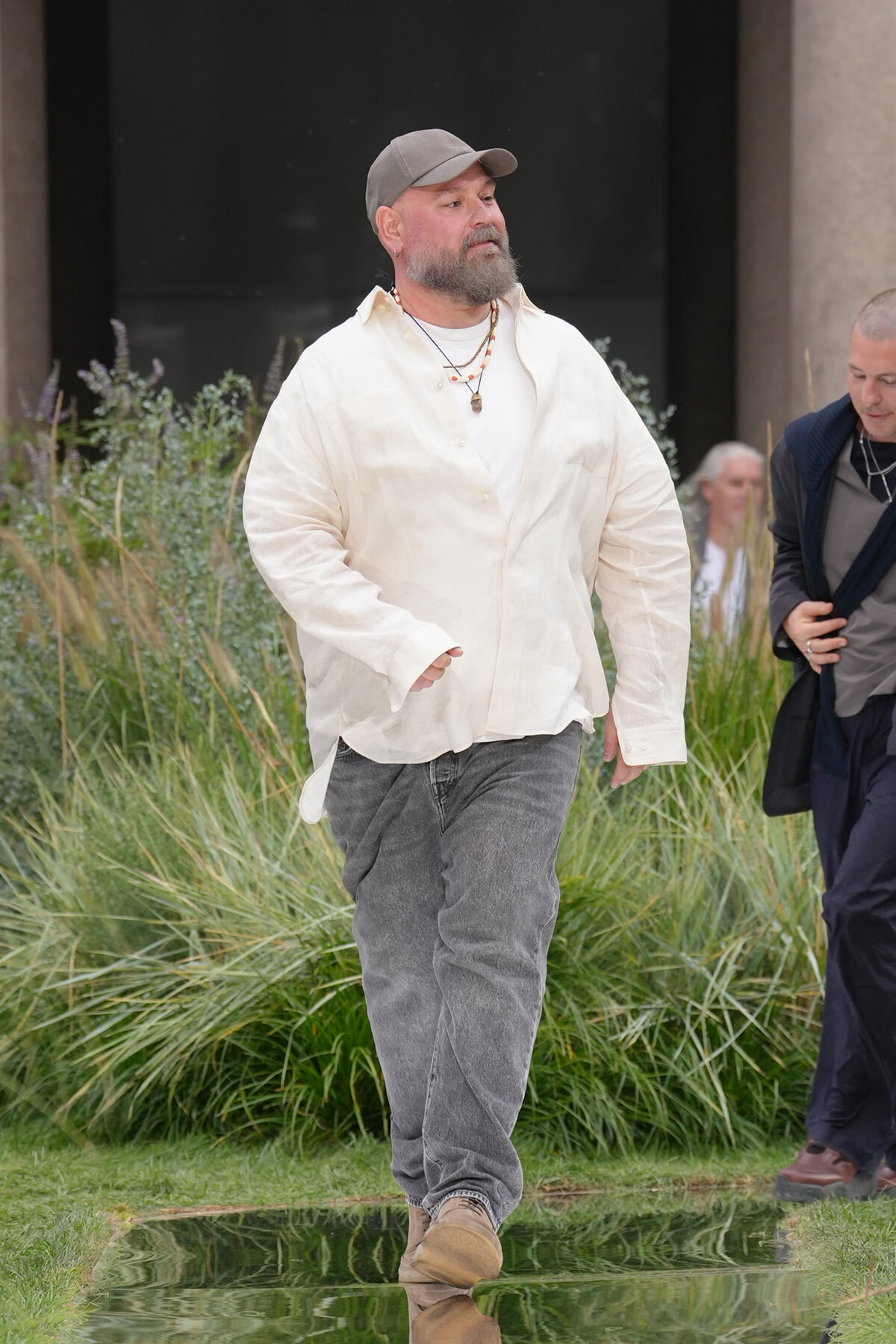 Bearded man in a light shirt and gray cap walking through a garden, with tall grasses in the background and a reflective puddle at his feet.