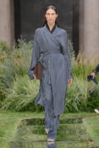 Female model walking a runway in a blue-gray wrap trench with belt, holding a brown clutch outdoors.
